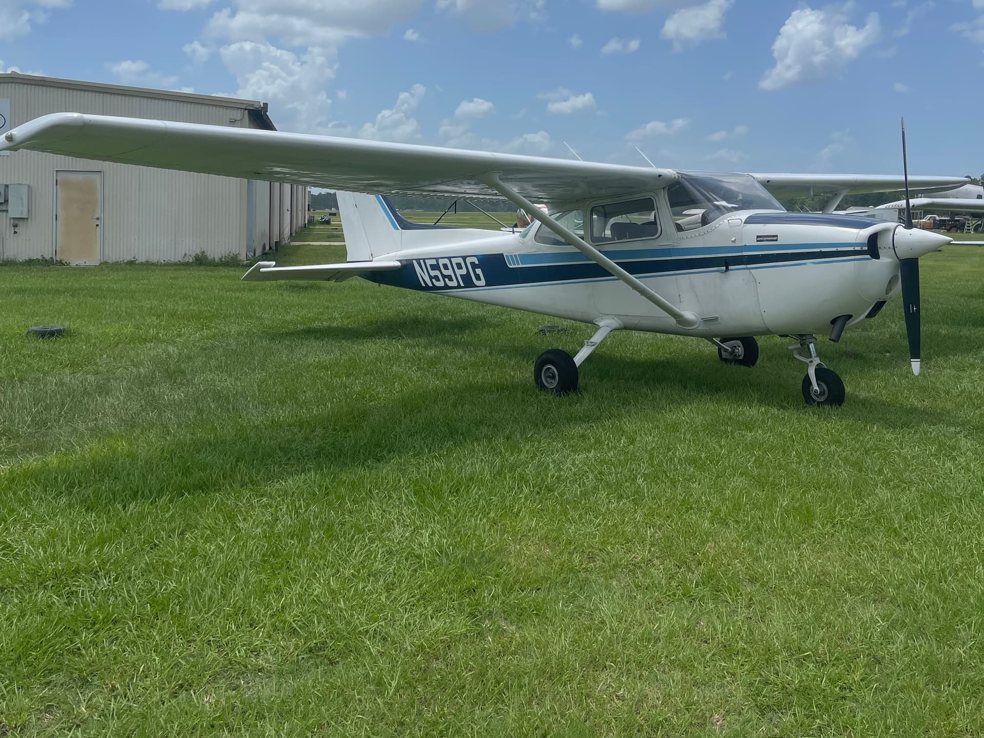 Cessna 172 N59PG, white with blue trim, parked on the grass beside a hangar